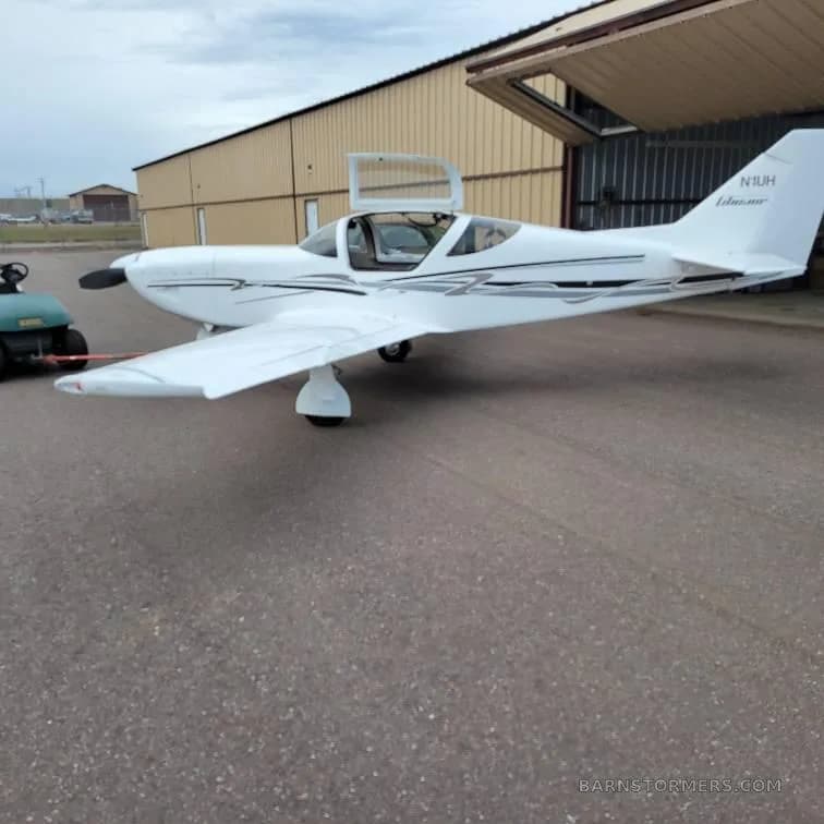 A white Vans RV-10 or similar kit plane with black and grey stripes, parked on an asphalt ramp with its canopy open, next to a hangar.