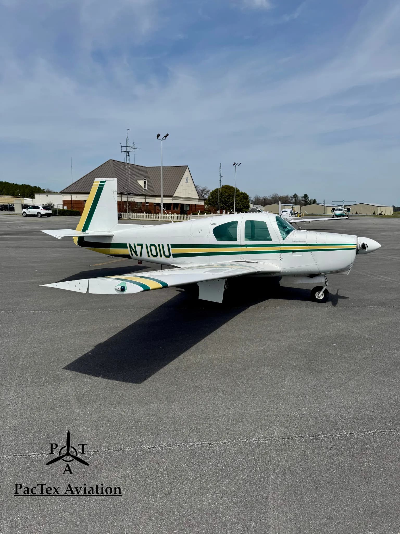 A white Mooney M20C aircraft with green and yellow stripes, parked on an airport tarmac with a building in the background.