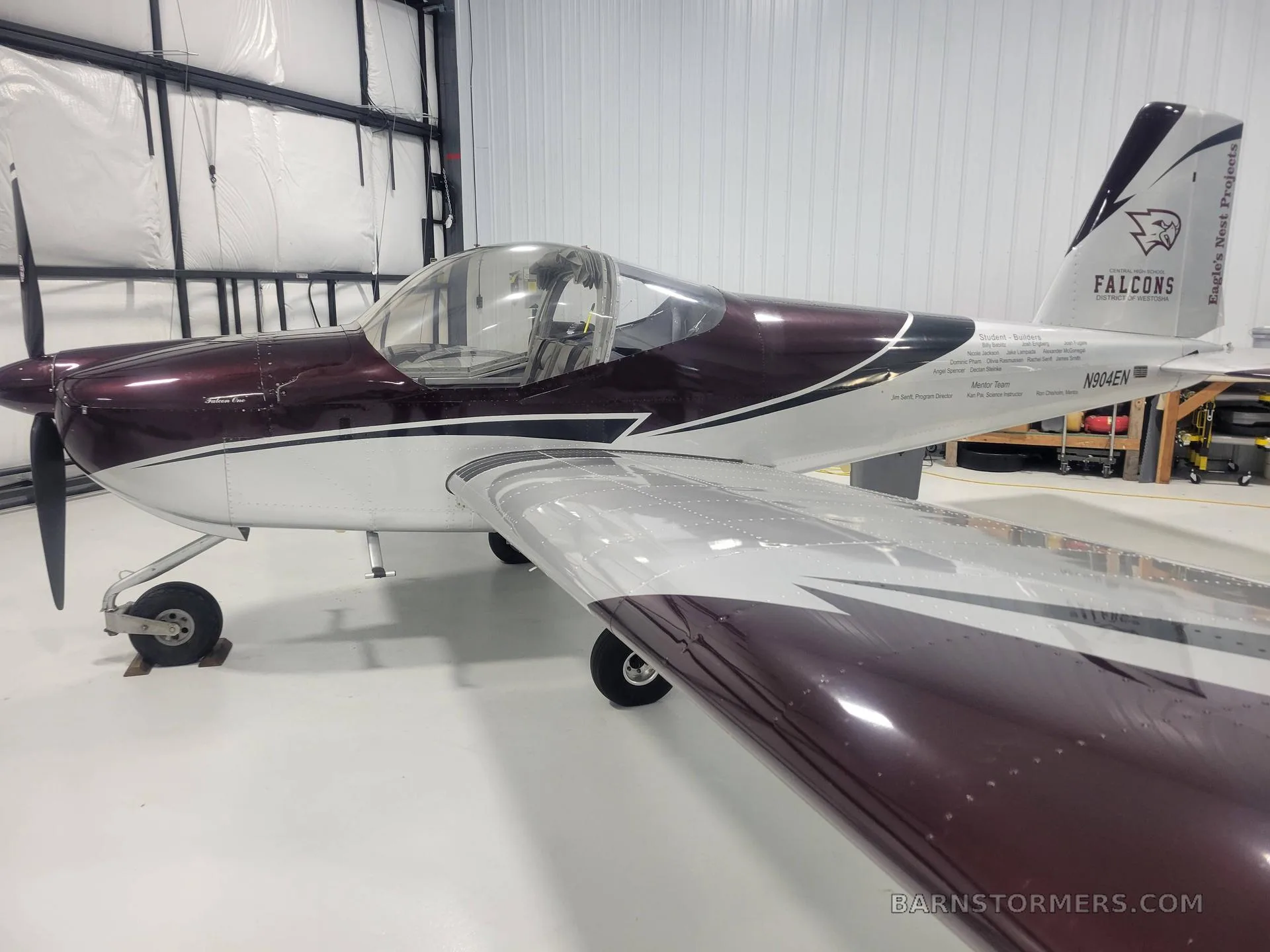 A burgundy and silver single-engine aircraft, N904EN, with a 'Falcons' logo on the tail, parked inside a white hangar.
