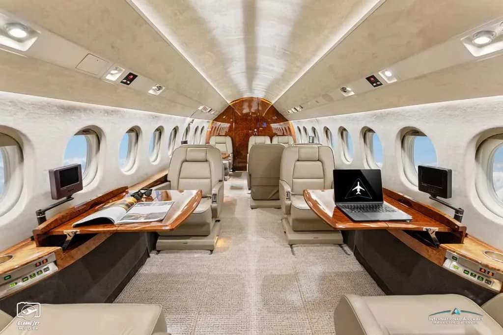 The luxurious cabin interior of a business jet, featuring beige leather seats, wooden tables, and a laptop on one table.