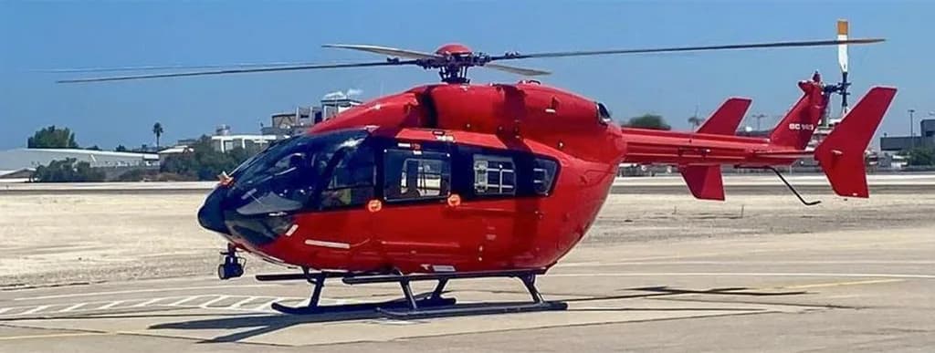 A red Airbus H135 helicopter with black skids and tinted windows parked on a sunny airfield.