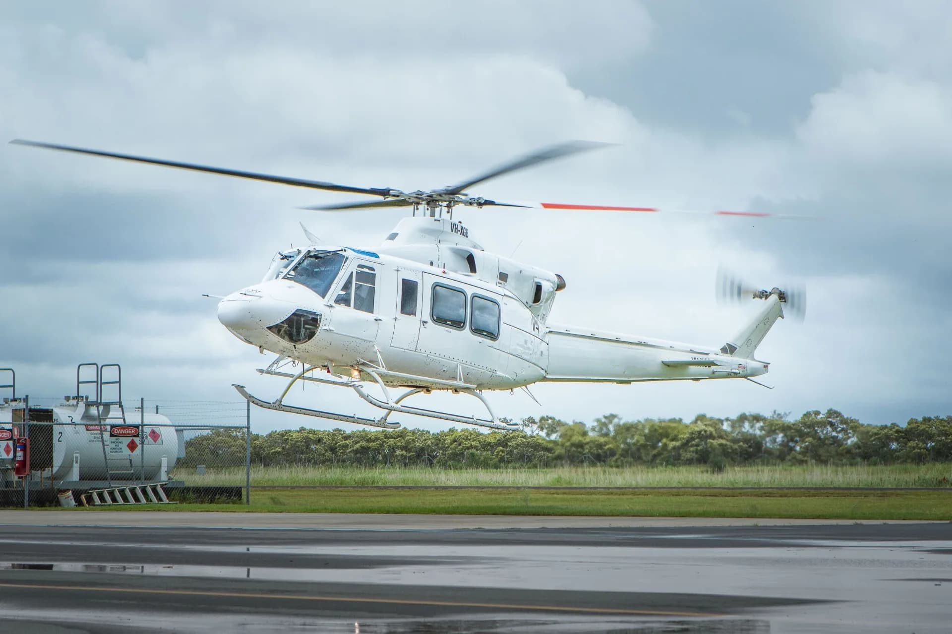 A white twin-engine helicopter, possibly a Bell 412, hovers just above a wet tarmac with fuel tanks and trees in the background under a cloudy sky.