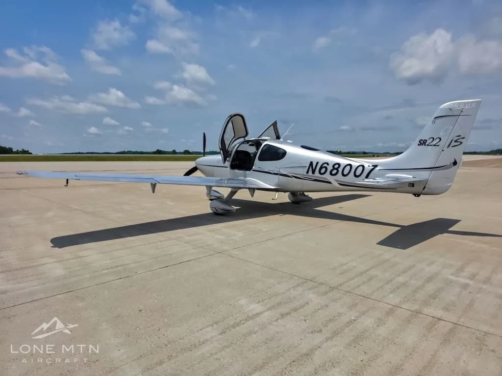 A silver Cirrus SR22 aircraft (N68007) with its gull-wing doors open, viewed from the rear-left on an airfield.