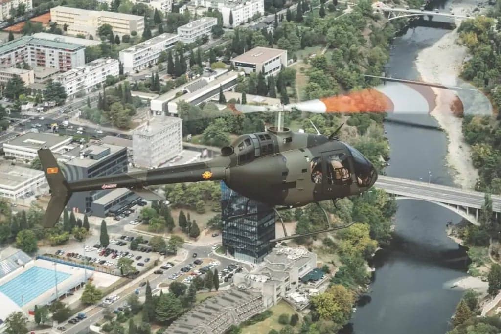 A camouflaged helicopter flies over a city and river.