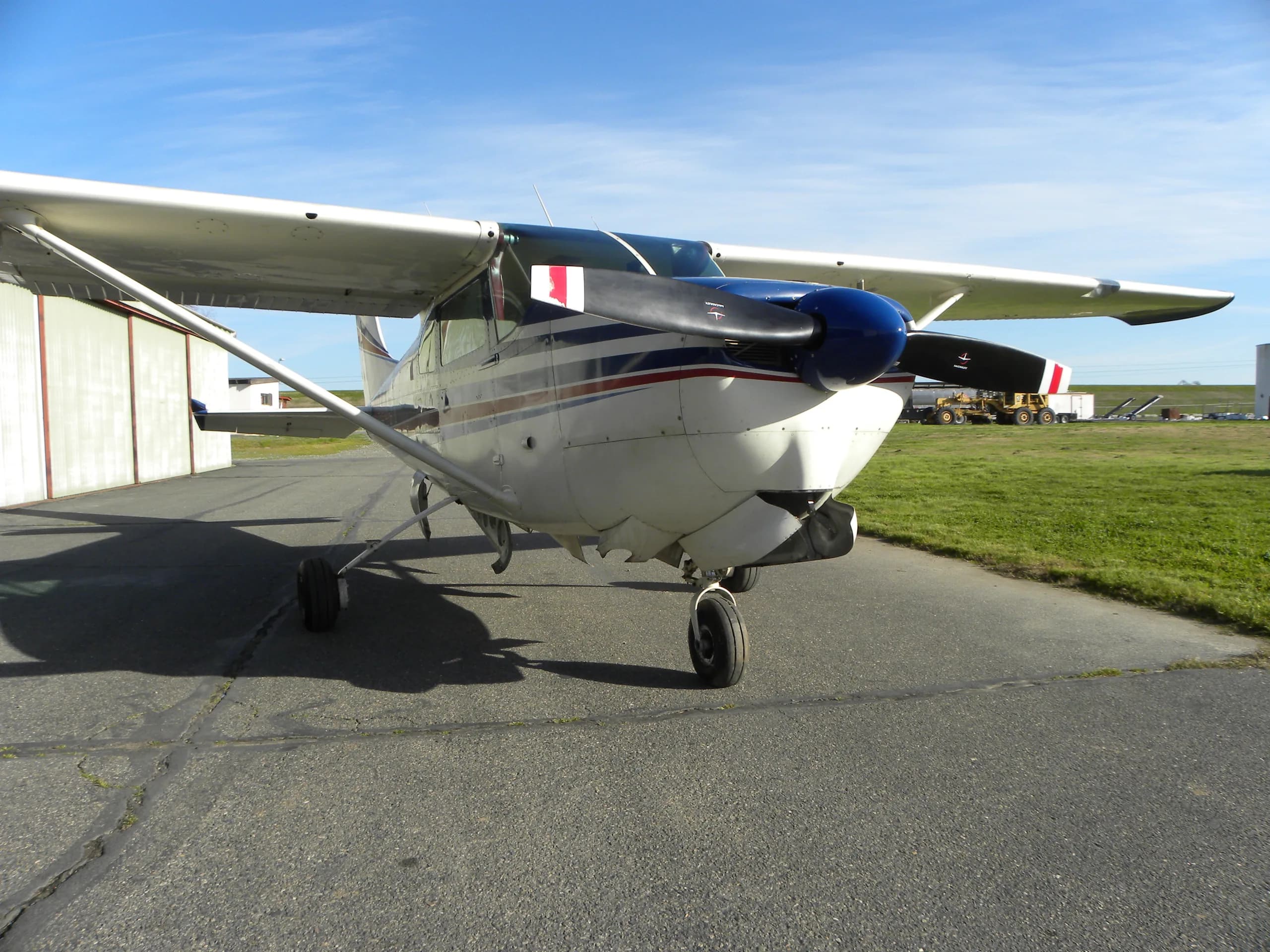 A white, blue, and red striped single-engine propeller aircraft with a blue spinner sits on an asphalt tarmac next to a hangar under a clear blue sky.