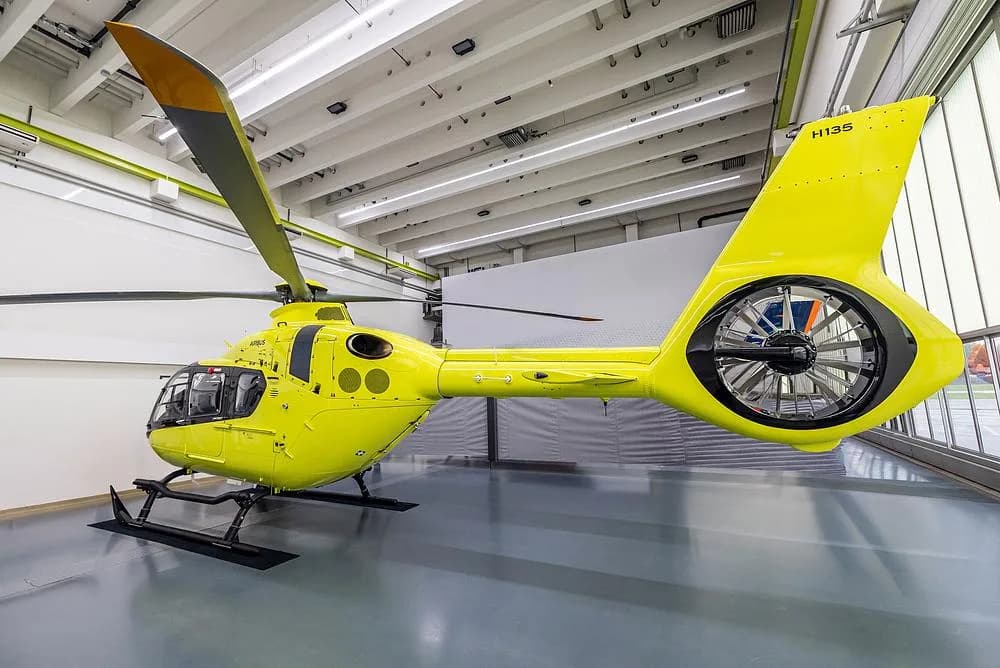 A bright yellow Airbus H135 helicopter is shown from its left side in a hangar, highlighting its Fenestron tail rotor.