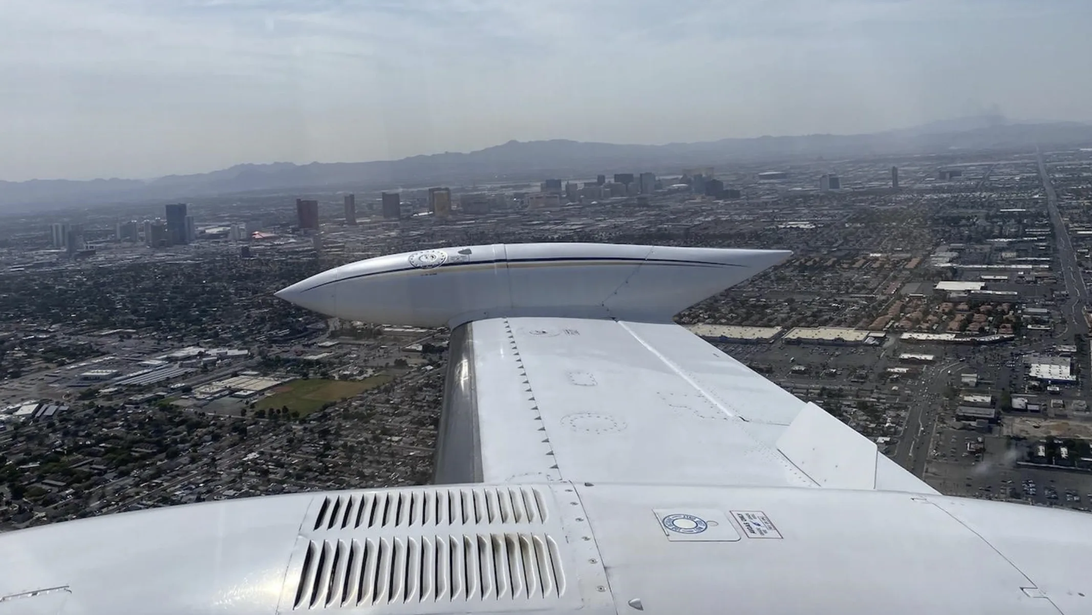 An aerial view from the wing of a white aircraft with blue and gold stripes, showing a city skyline in the distance under a hazy sky.