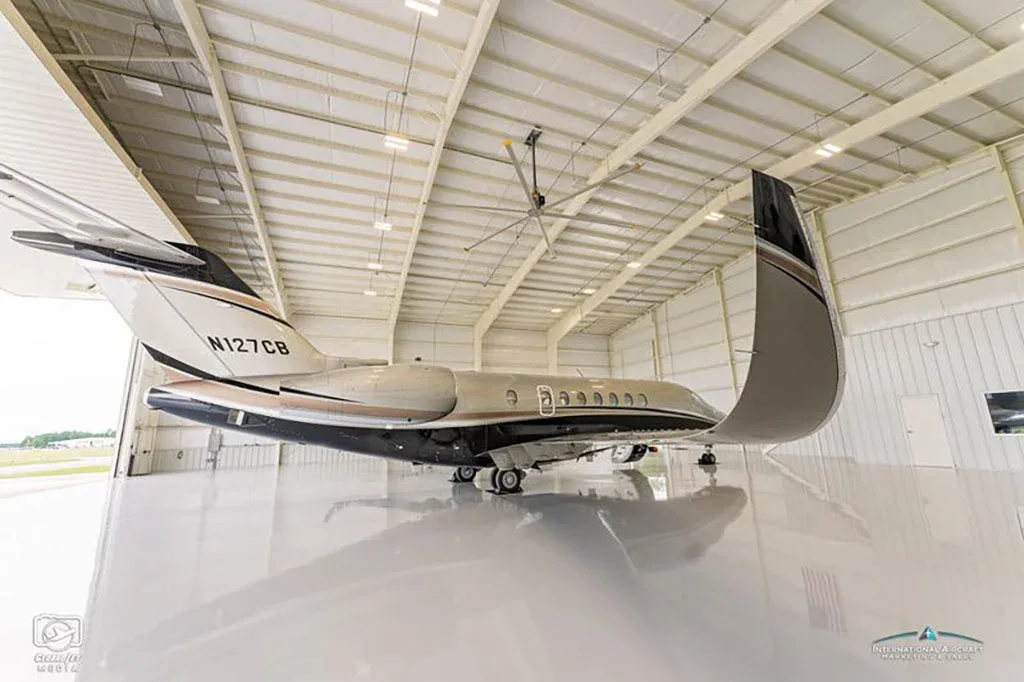 A rear-quarter view of a grey and black business jet with registration N127CB parked inside a hangar.