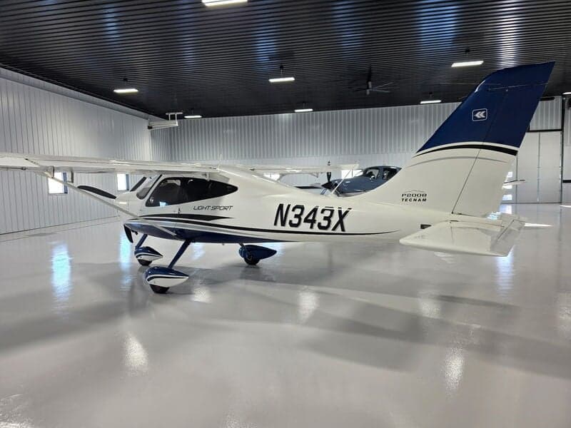 A white and blue Tecnam P2008 Light Sport aircraft (N343X) is parked in a hangar, showing the tail with a blue stripe and logo.