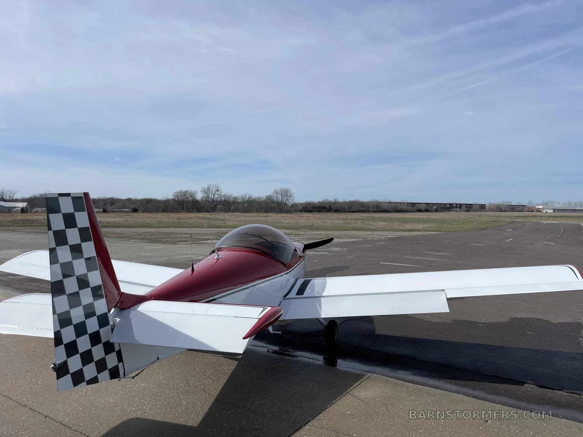 A rear-left view of a red and white single-engine aircraft with a checkered tail, parked on an apron next to a runway.