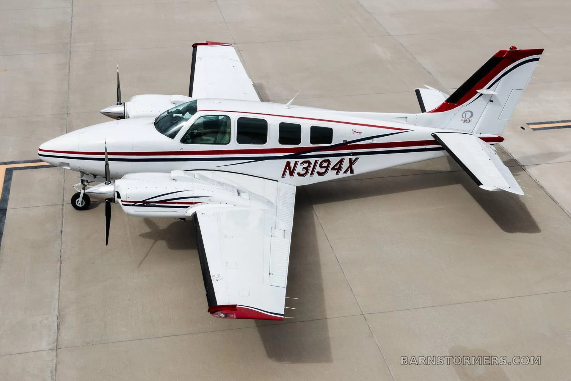 An overhead view of a white twin-engine propeller aircraft with red and blue stripes, registration N3194X, parked on concrete.