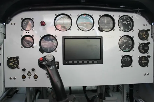 An aircraft cockpit instrument panel displaying multiple analog gauges, one digital screen, and a control stick.