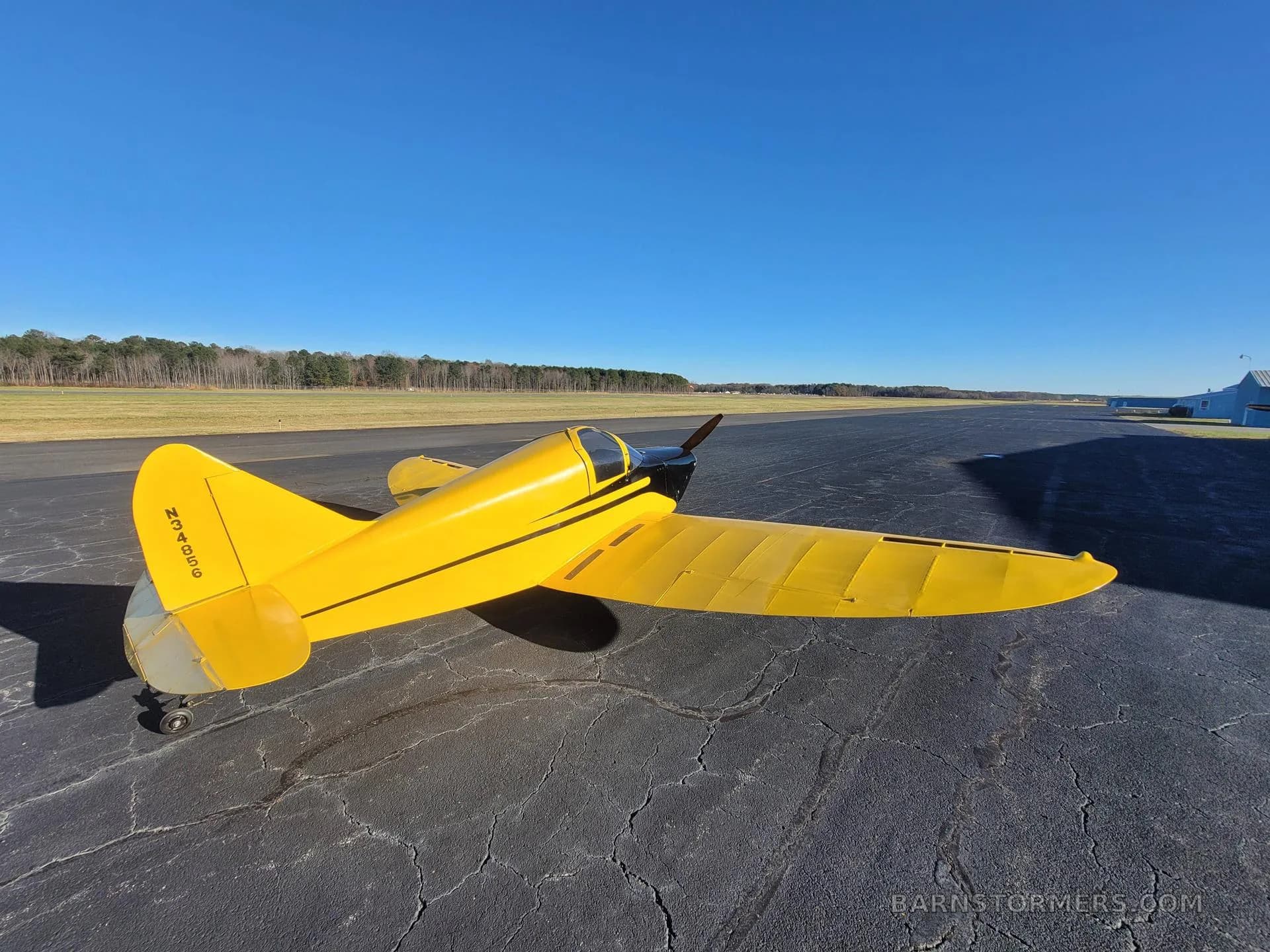 A yellow single-engine aircraft with black accents, viewed from the rear-left, on an asphalt runway with a building in the distance.