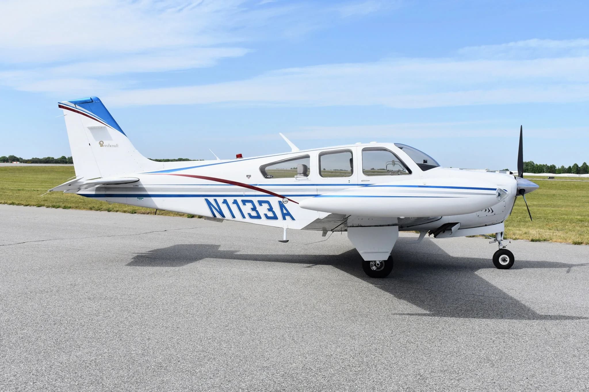 A white Beechcraft Bonanza F33A with blue and maroon stripes, registration N1133A, parked on an asphalt tarmac.