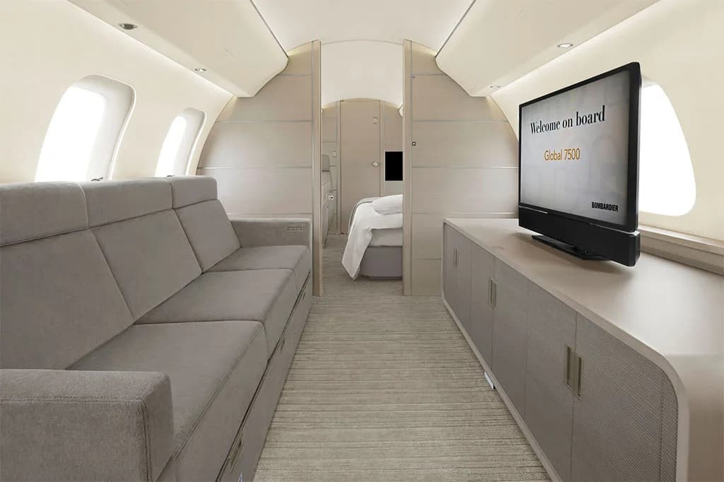 A private jet lounge area with a grey sofa, a large television displaying "Welcome on board Global 7500", and a view into a bedroom.