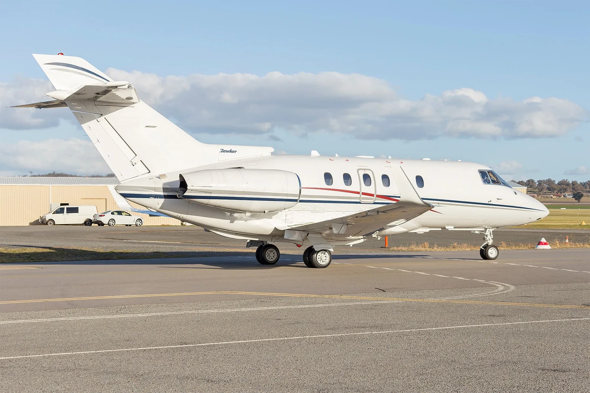 A white private jet with blue and red stripes, labeled "Hawker", parked on an airfield on a sunny day.