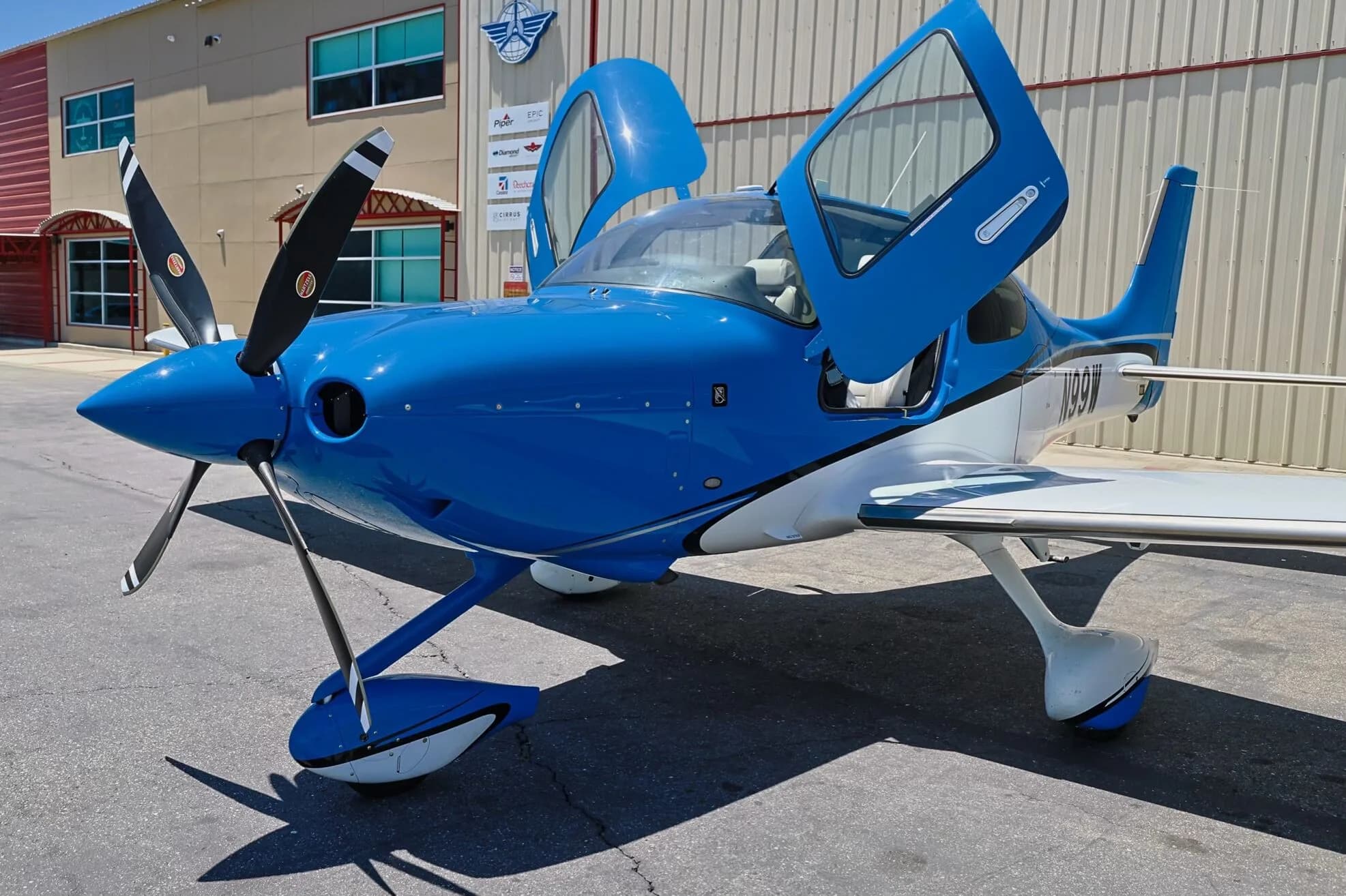 Front three-quarter view of a blue and white Cirrus SR22 aircraft (N99W) with open doors, parked next to a building.