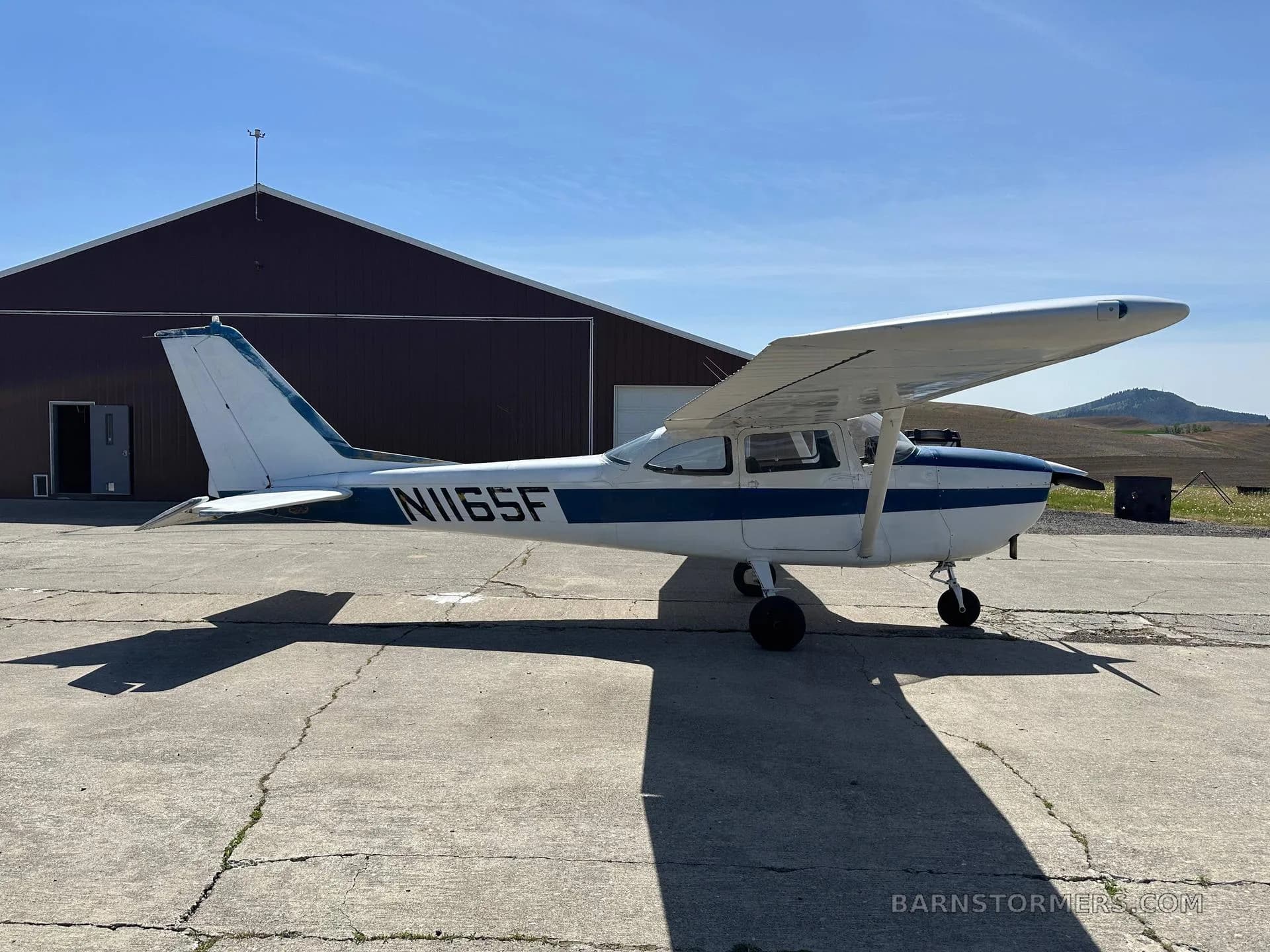 A white and blue Cessna N1165F aircraft is parked sideways on a concrete apron next to a red hangar.