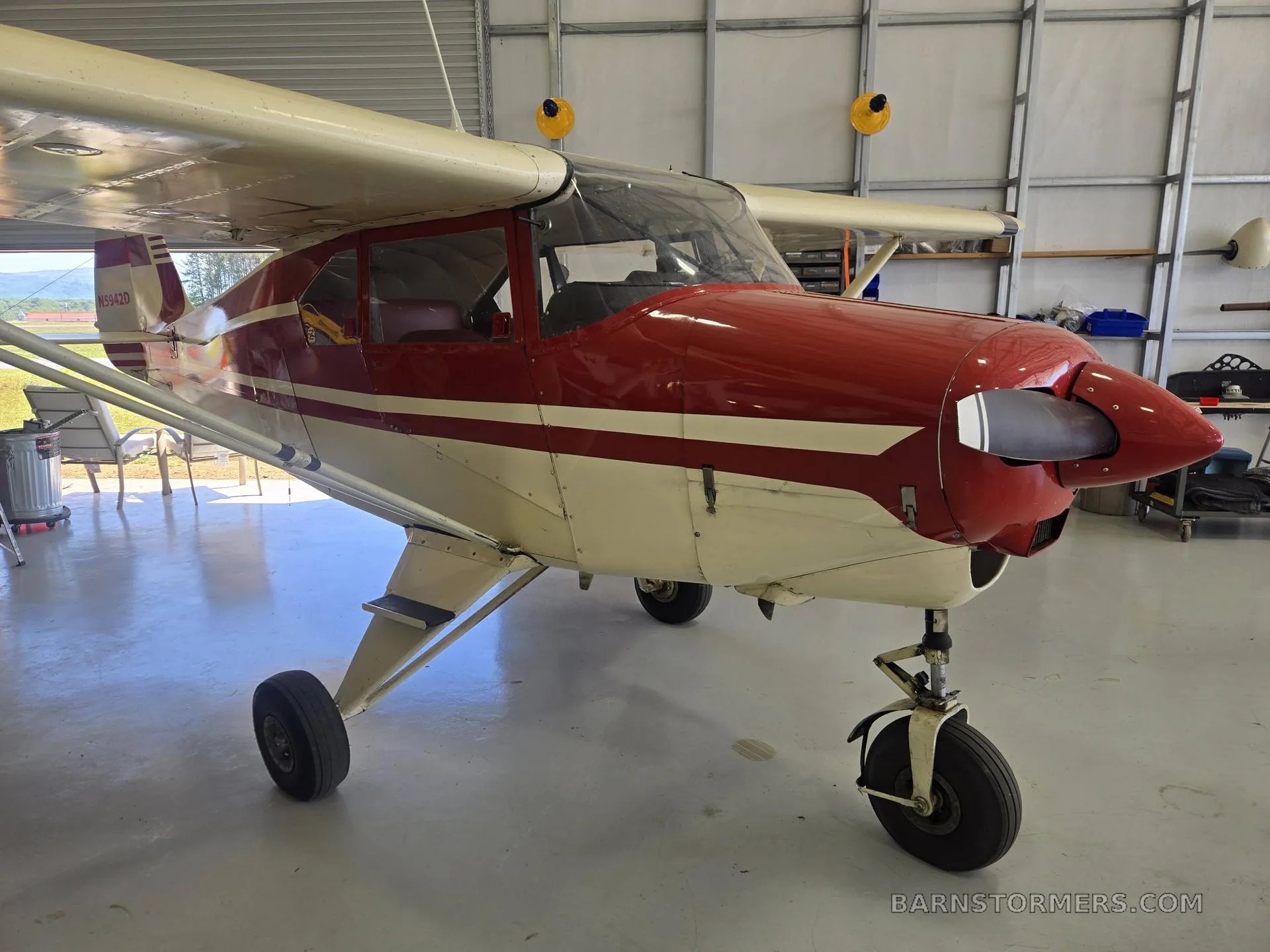 A red and white single-engine propeller aircraft with tricycle landing gear, parked in a hangar, viewed from the front-right.