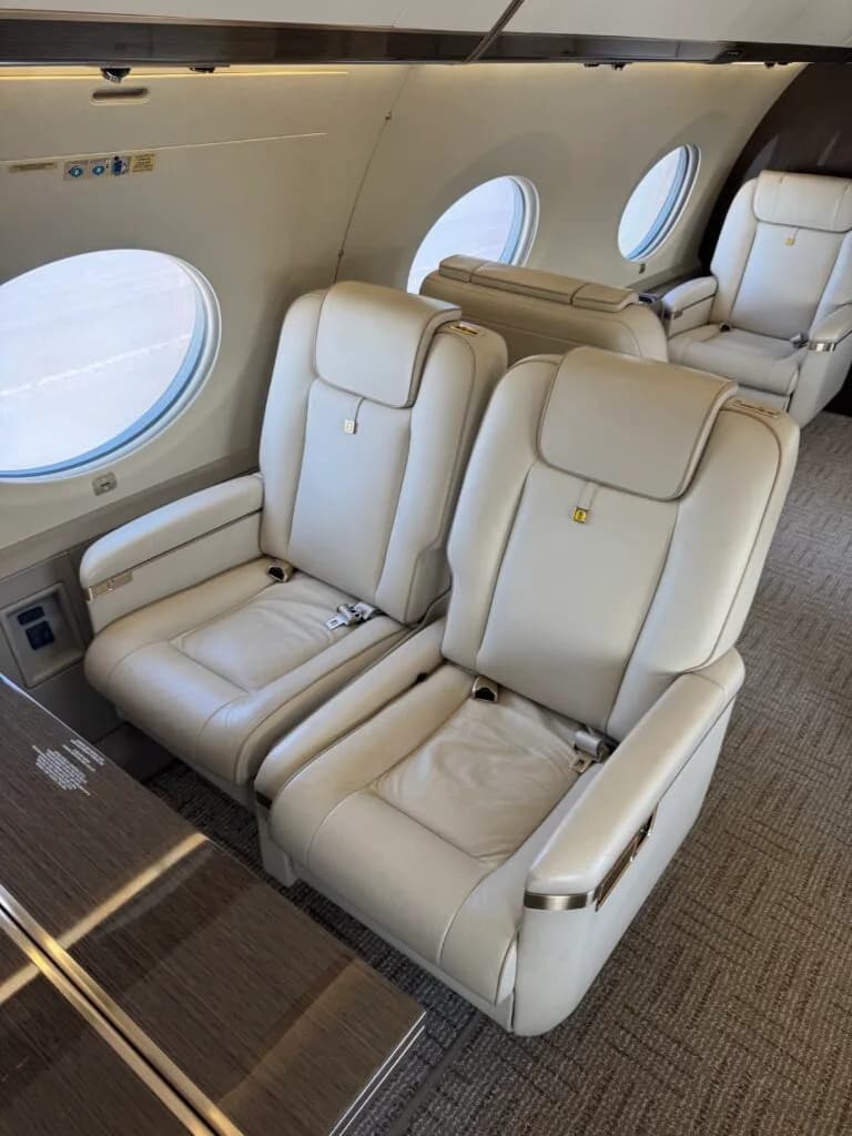 Two light-colored leather seats in the Gulfstream cabin, positioned next to a window and a dark wood table.