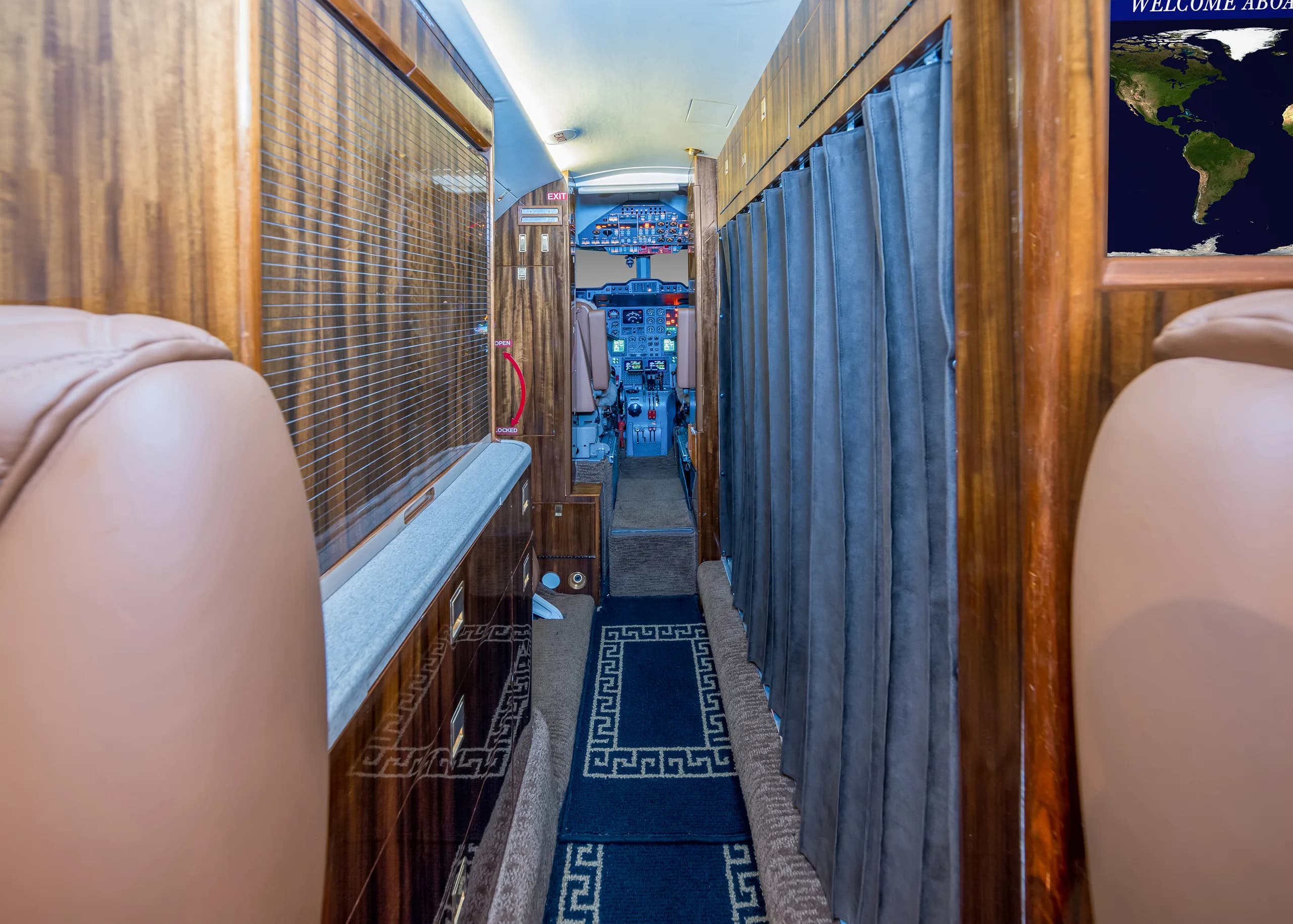A view down the aisle of a private jet cabin towards the cockpit, with wood paneling and blue curtains.