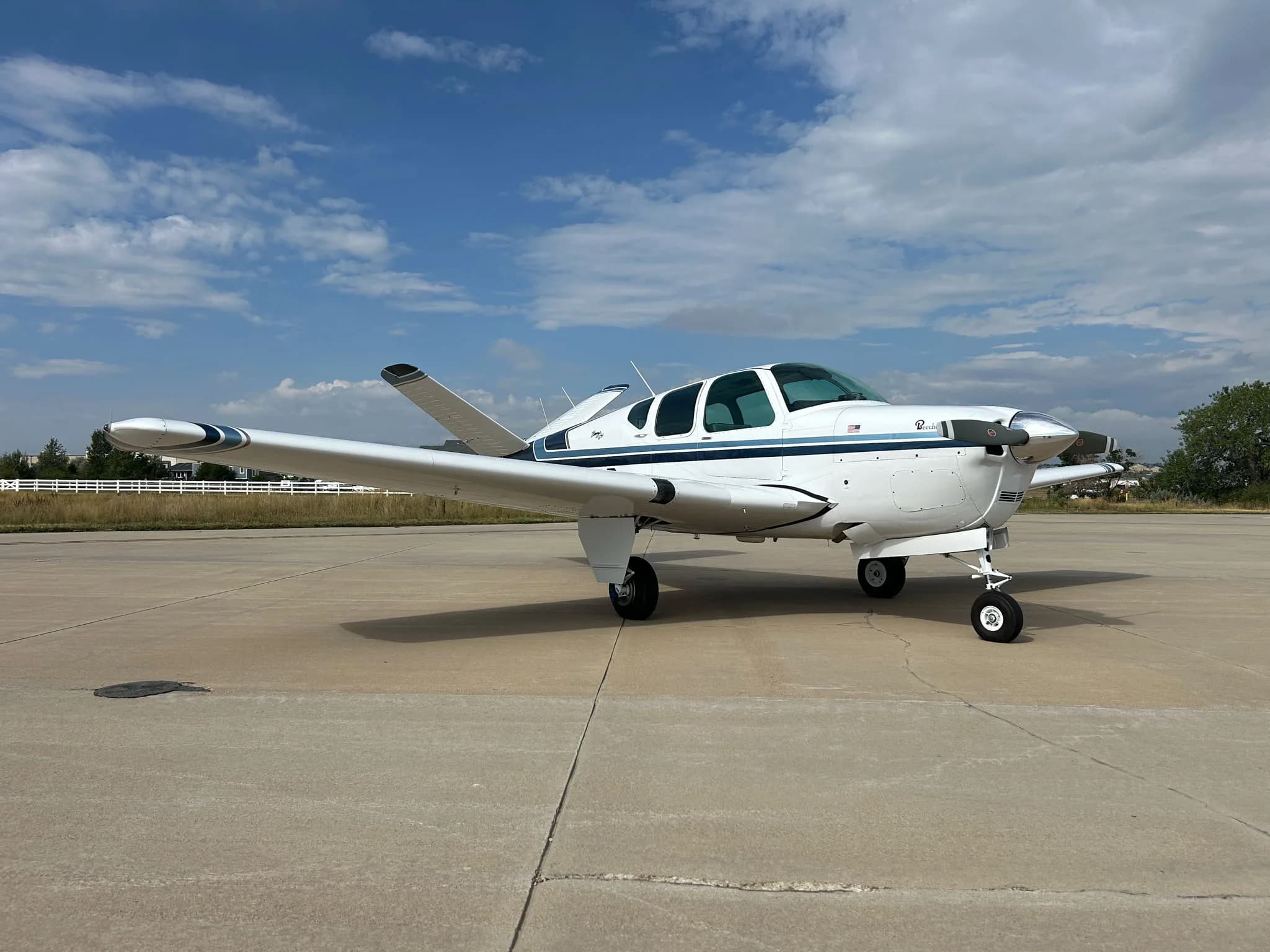 A white Beechcraft Bonanza with blue stripes parked on a concrete tarmac under a partly cloudy sky.