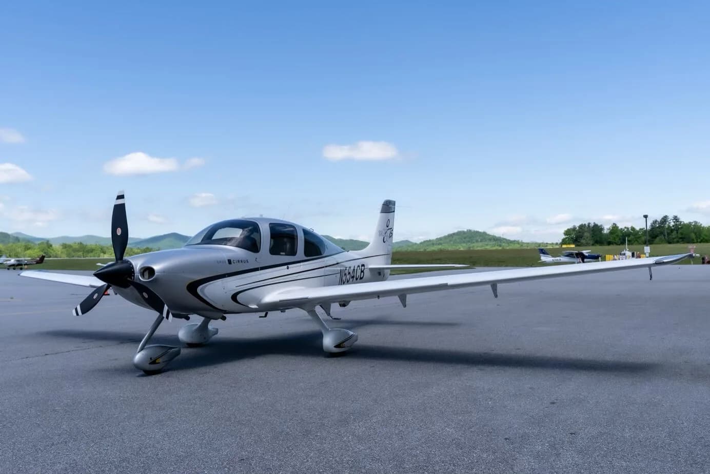 A silver and white Cirrus SR22T aircraft with registration N554CB parked on an airport tarmac with mountains in the background.