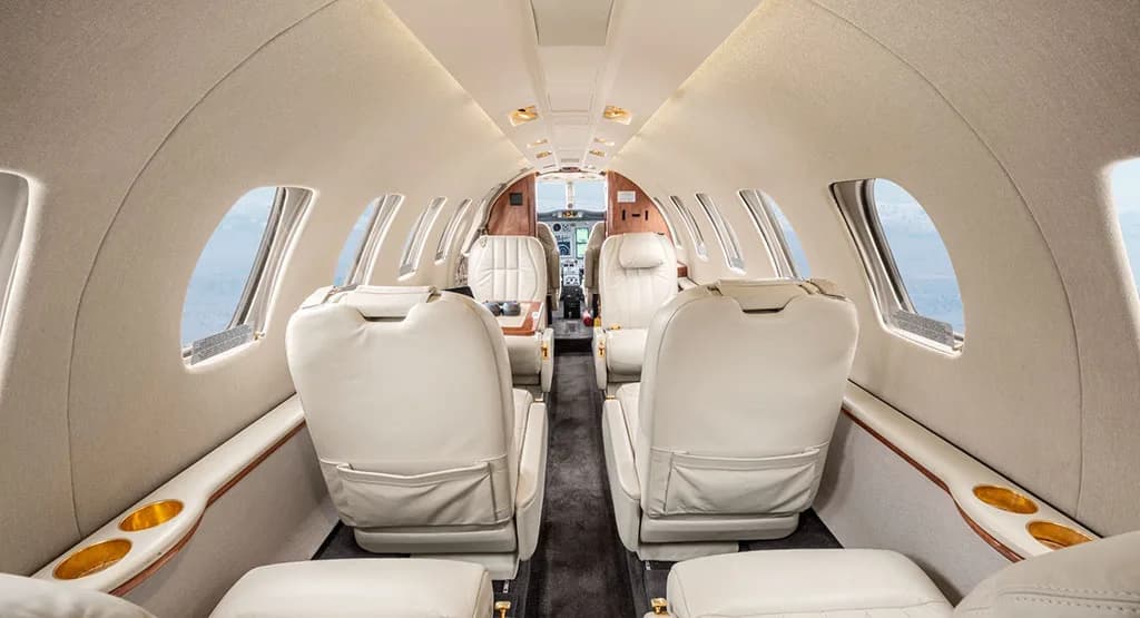 The aircraft's cabin interior features a central aisle, light cream leather seats, wooden accents, and a view towards the cockpit.