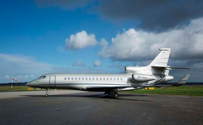 A silver Dassault Falcon business jet with registration OY-OLU is parked on a tarmac under a blue sky with scattered clouds.
