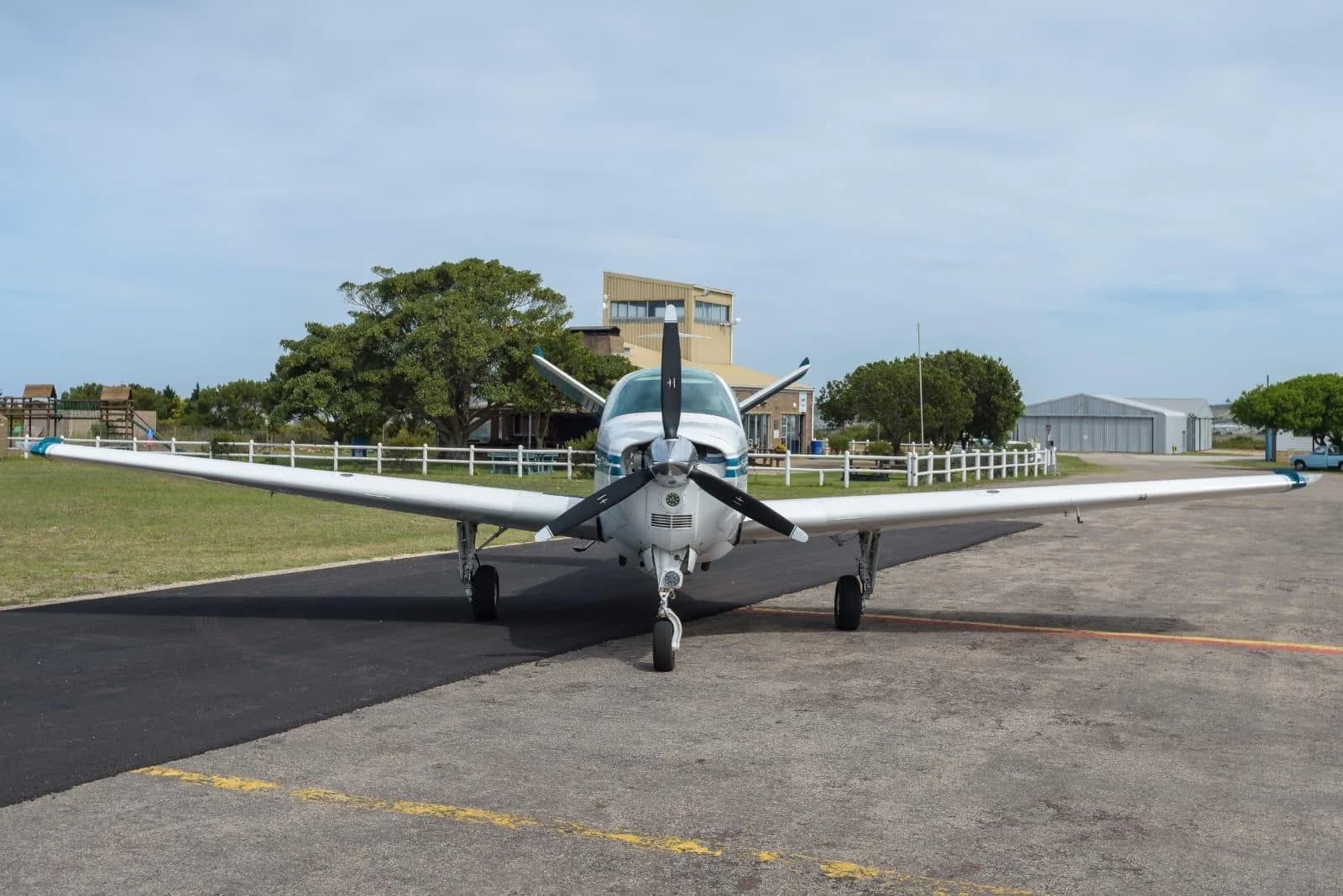 A white single-engine propeller aircraft with blue stripes is parked on an asphalt tarmac at an airfield, viewed from the front.