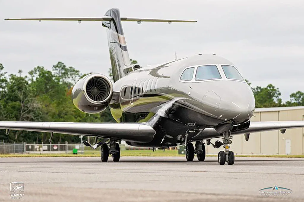 A front-quarter view of a grey and black business jet with a gold stripe on a tarmac under a cloudy sky.