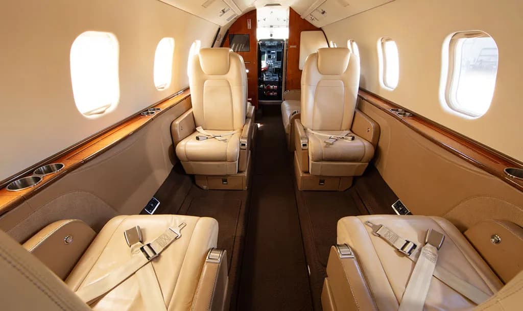 The aircraft cabin interior with light tan leather seats and wood trim, looking towards the cockpit.