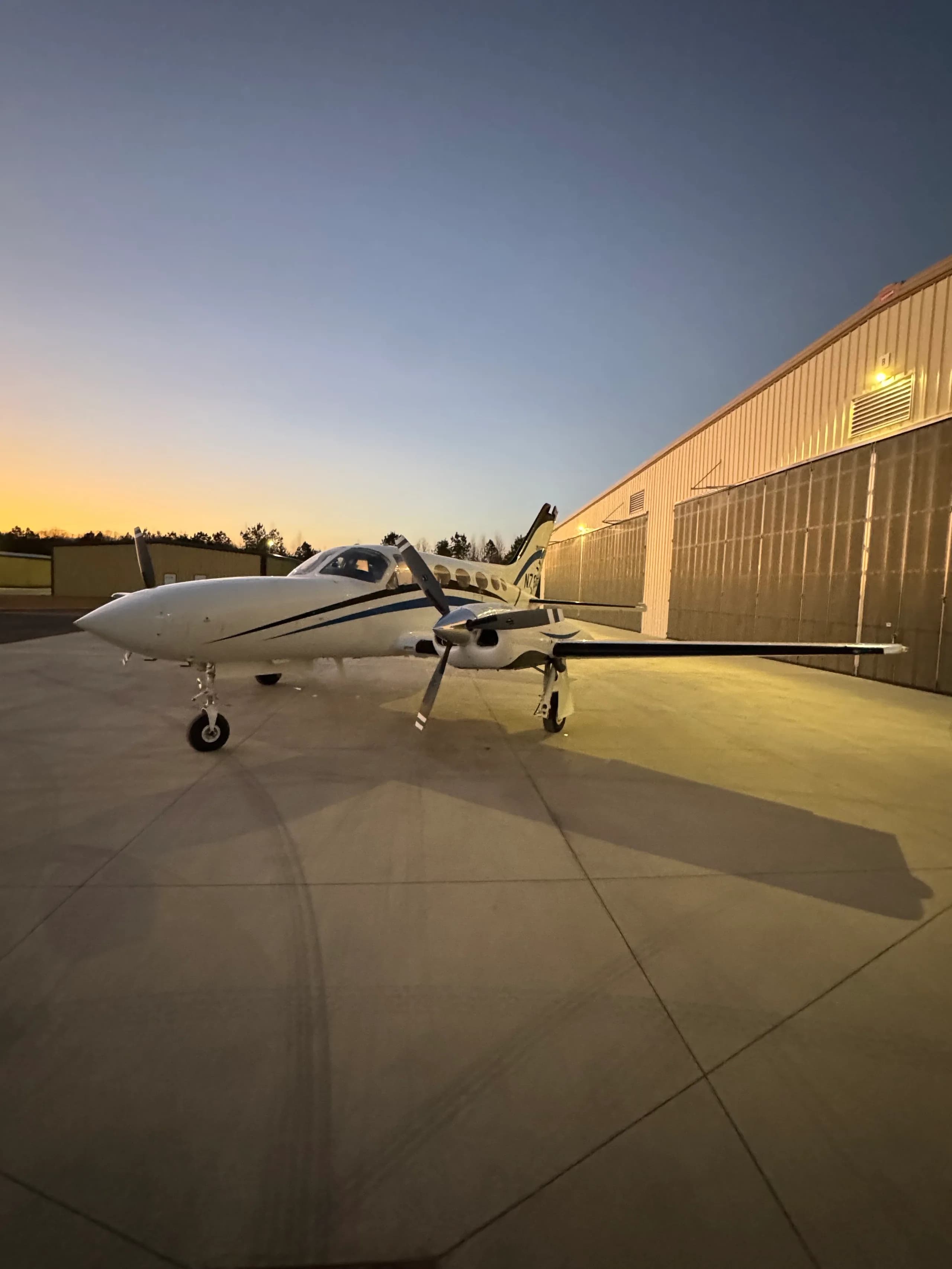 A white twin-engine aircraft with blue stripes, N762CA, parked on an airfield with a hangar in the background.
