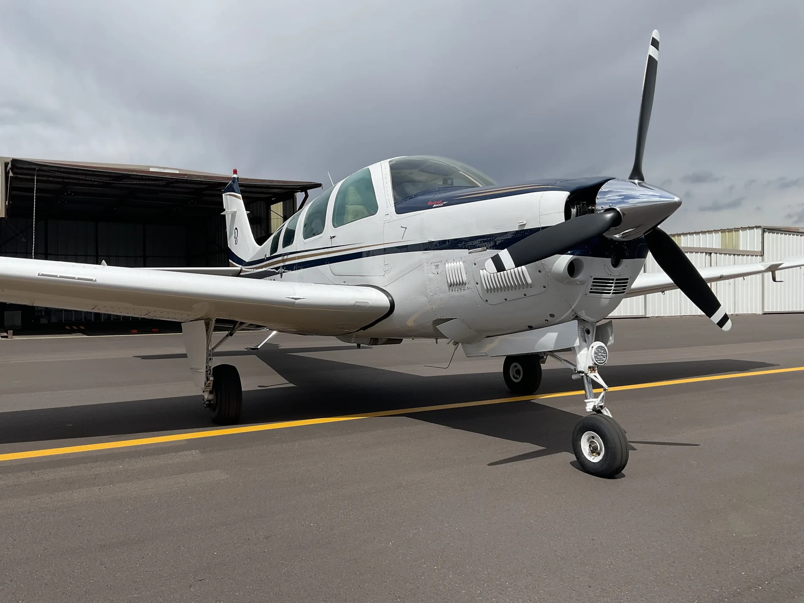 A white and blue Beechcraft Bonanza with a V-tail is parked on an asphalt tarmac under a cloudy sky.
