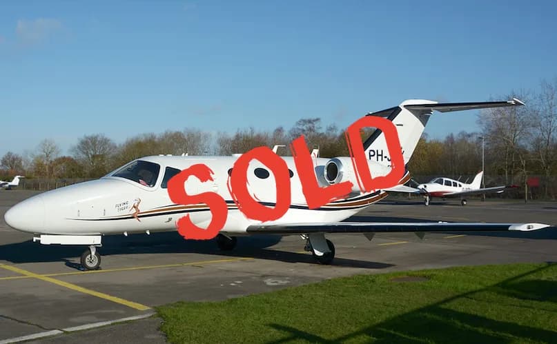A white private jet with brown and black stripes, featuring 'Flying Tiger' text and a pin-up graphic, is parked on an airfield under a clear blue sky.