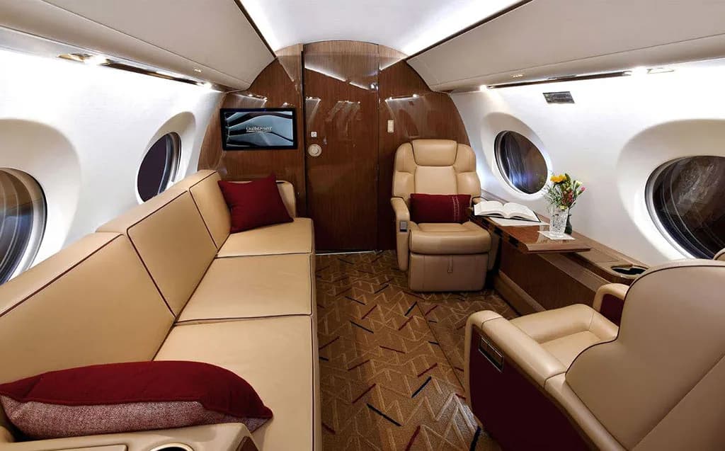 The aft section of a private jet cabin with a long beige sofa, a single seat, and a small table with flowers.