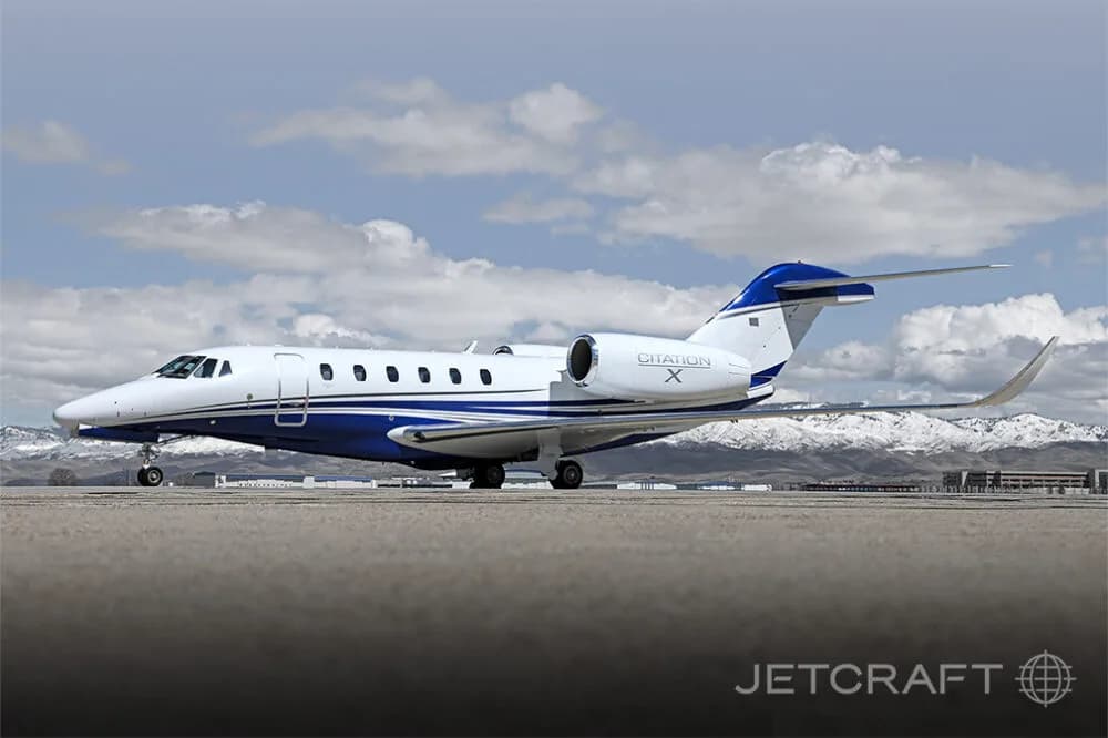 A white and blue Cessna Citation X business jet is parked on an airfield with snow-capped mountains in the background.