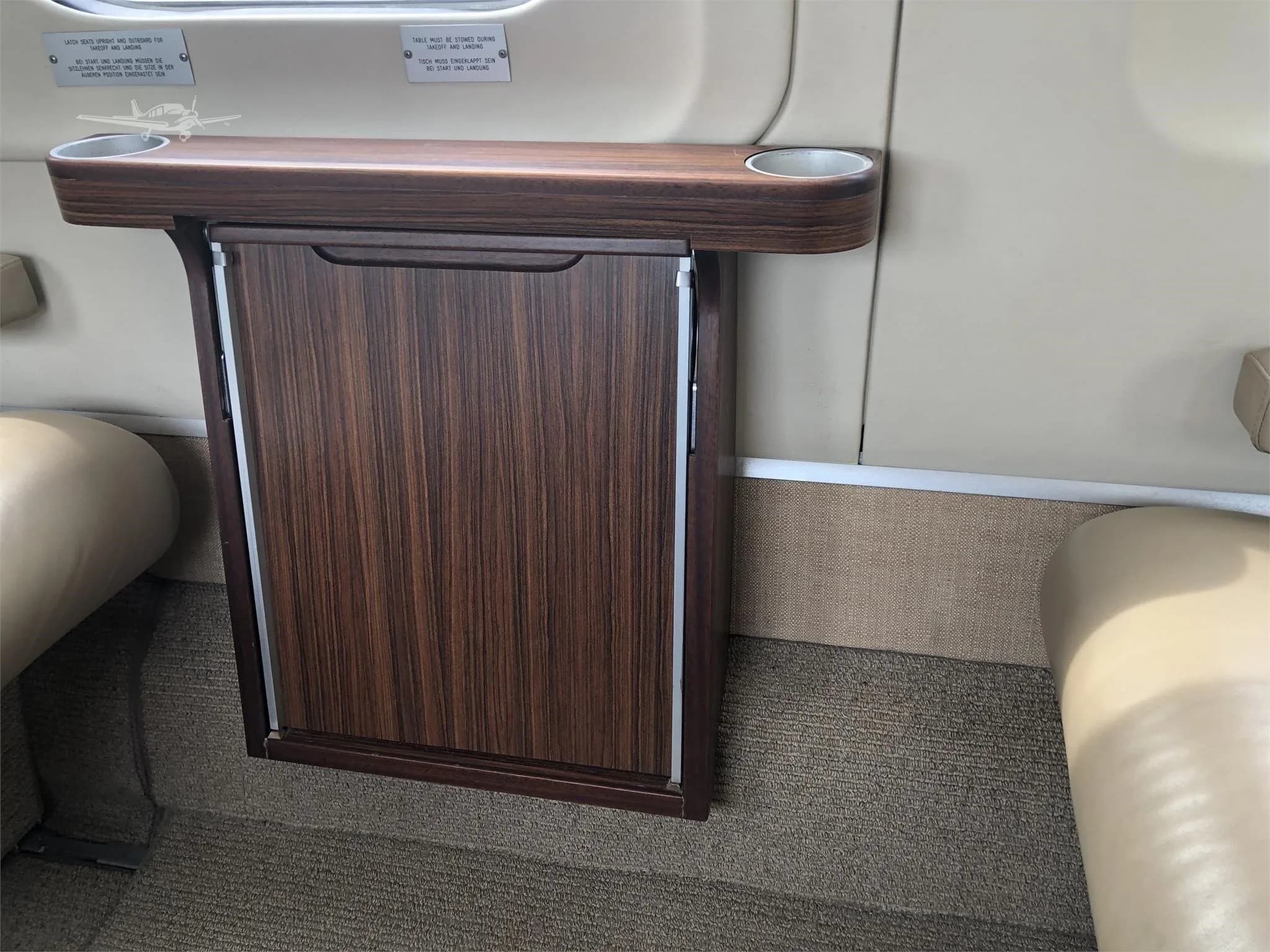 A view of a private jet's cabin interior, showing a wooden fold-out table with cup holders between two beige leather seats.