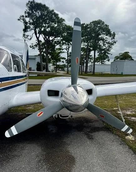 A close-up view of the shiny propeller spinner and gray blades of a twin-engine aircraft, with trees and hangars in the background.