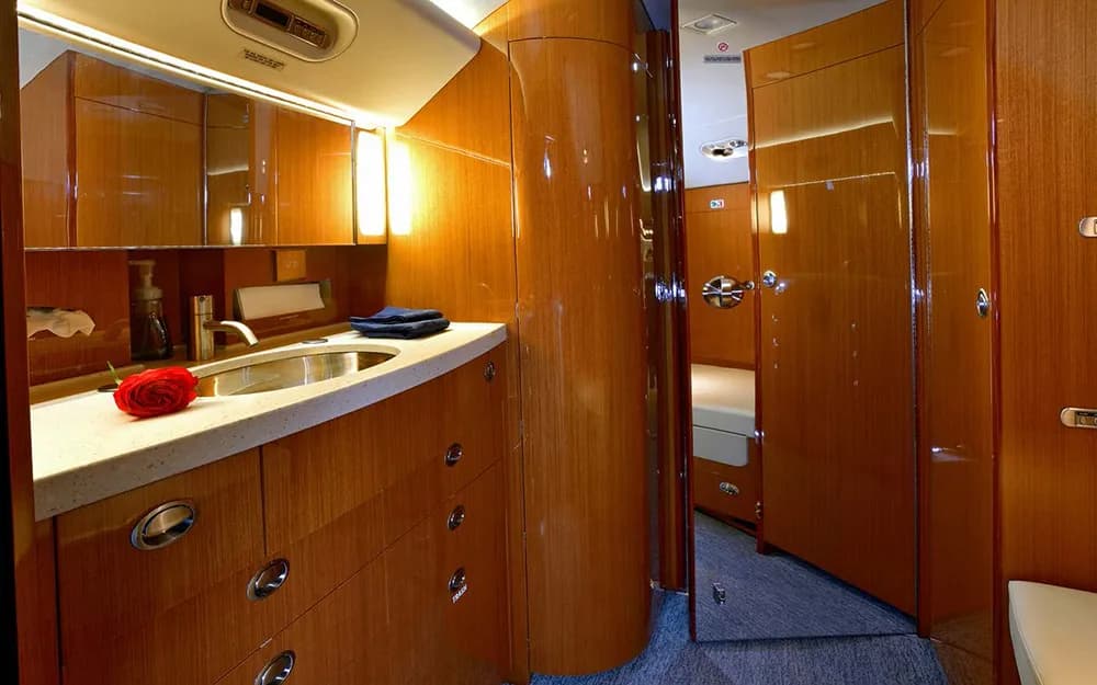The aircraft's lavatory with a modern sink, mirror, wooden cabinetry, and a red rose, with a sleeping berth visible beyond.
