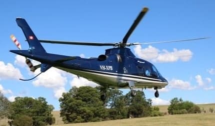 A blue Agusta Grand helicopter taking off from a grassy field.