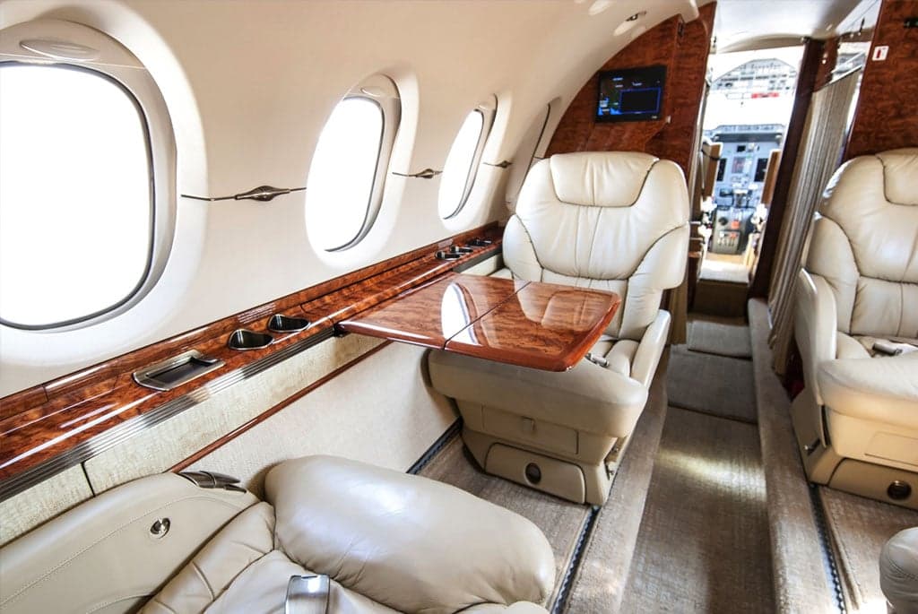 The luxurious interior of a business jet cabin, featuring cream leather seats, a polished wood table, and large windows.