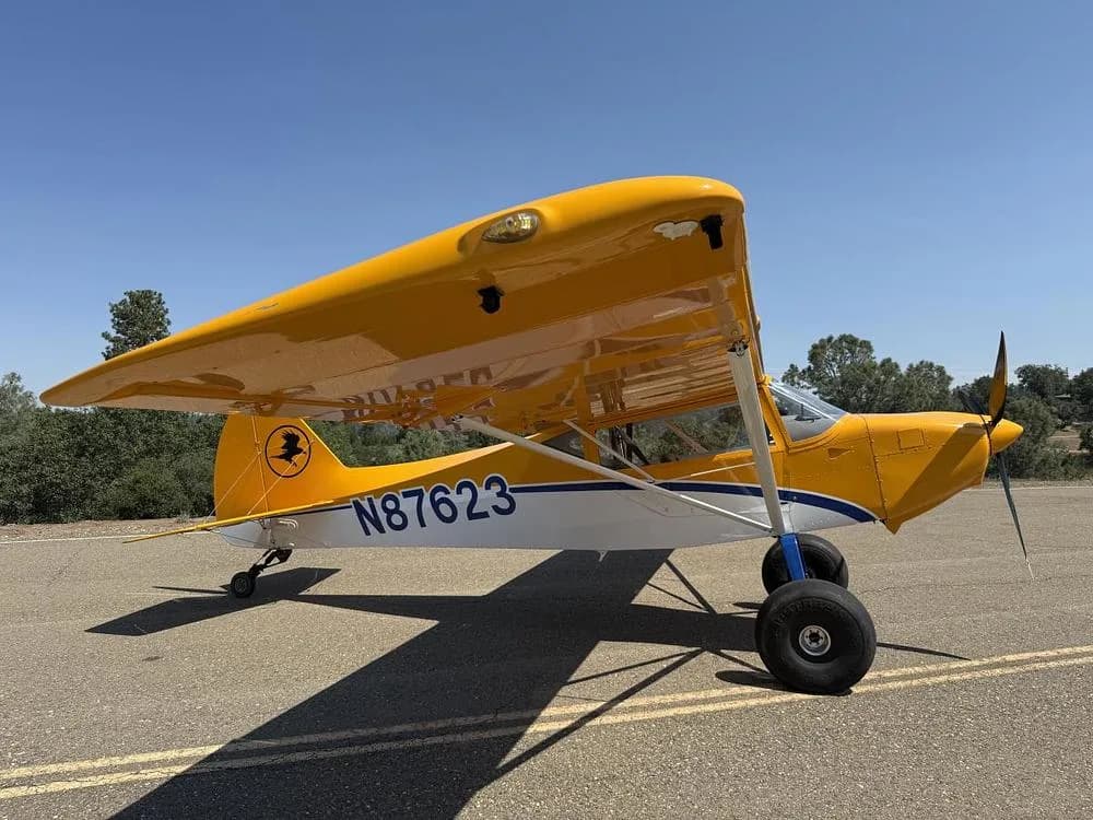 A yellow and white high-wing aircraft, N87623, with large tires, is parked on an asphalt runway under a clear blue sky, viewed from the front-right.