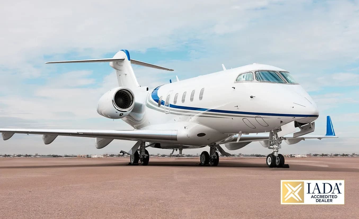 A white business jet with blue and grey stripes is parked on an airfield under a partly cloudy sky.
