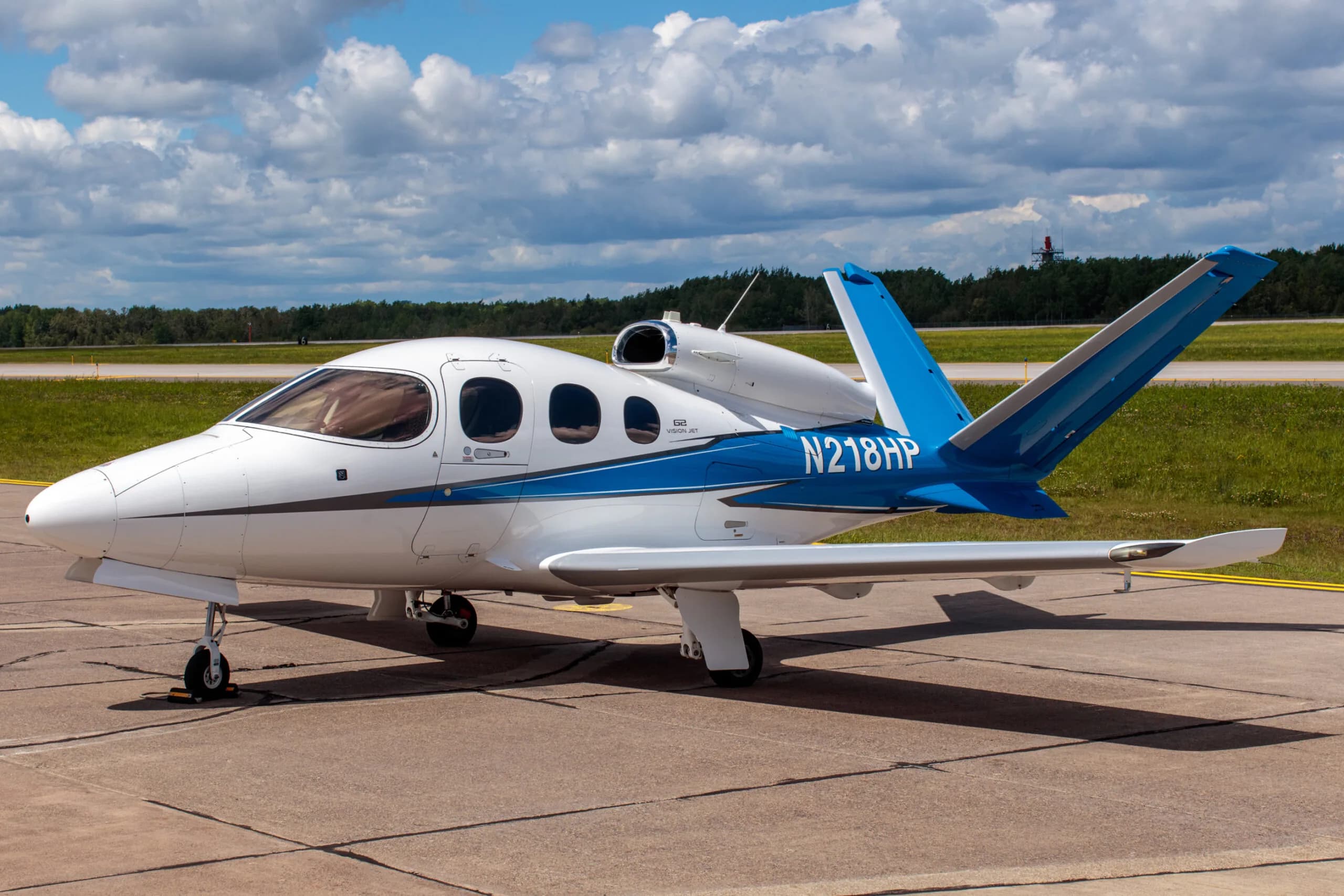 A white and blue Cirrus Vision Jet (N218HP) parked on a tarmac under a partly cloudy sky.