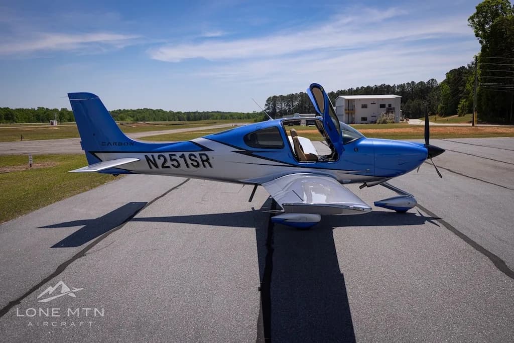 A side view of the blue and silver Cirrus SR20 aircraft (N251SR) with open doors, parked on an airfield with buildings in the background.