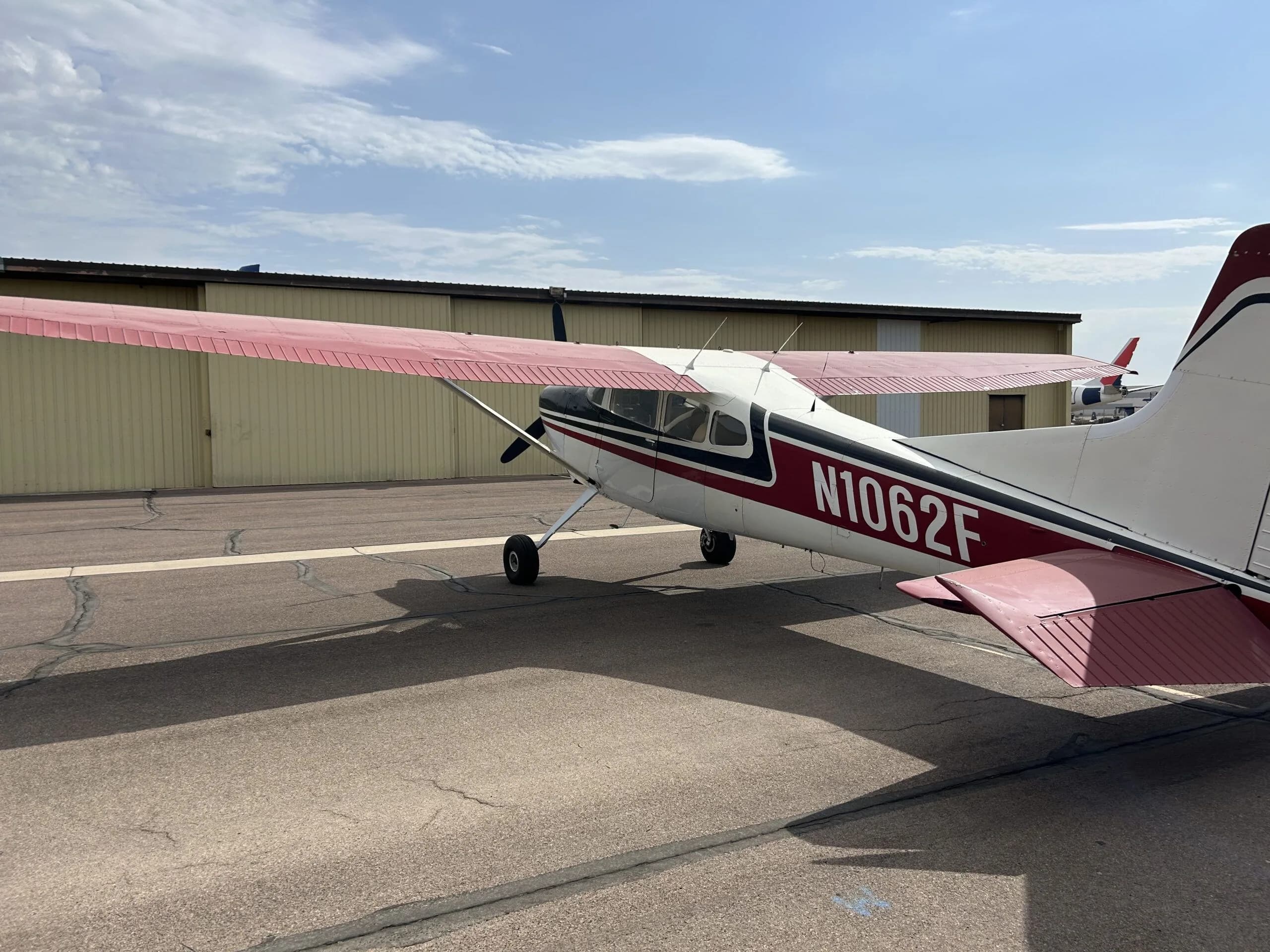 A white, black, and red Cessna 180 aircraft (N1062F) parked on an airport tarmac, viewed from the rear-left.
