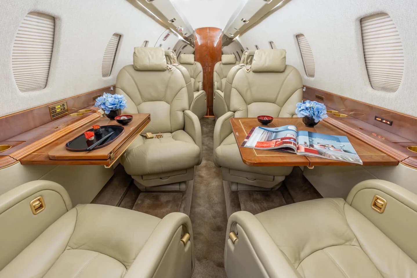 The luxurious interior cabin of a business jet with beige leather seats, wooden tables, and blue floral arrangements.