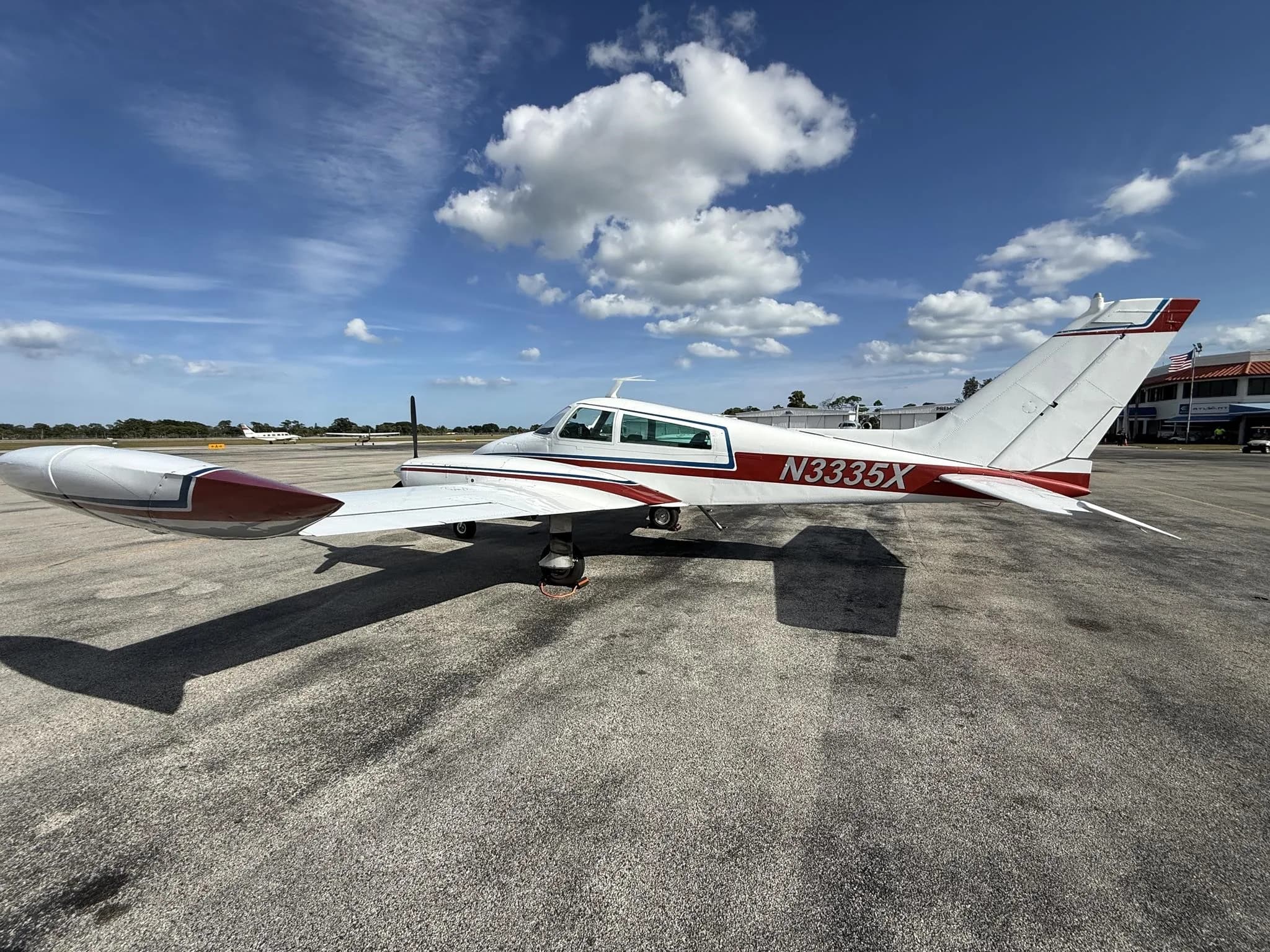 A white Cessna 310L with red and blue stripes parked on an airport tarmac under a partly cloudy sky.