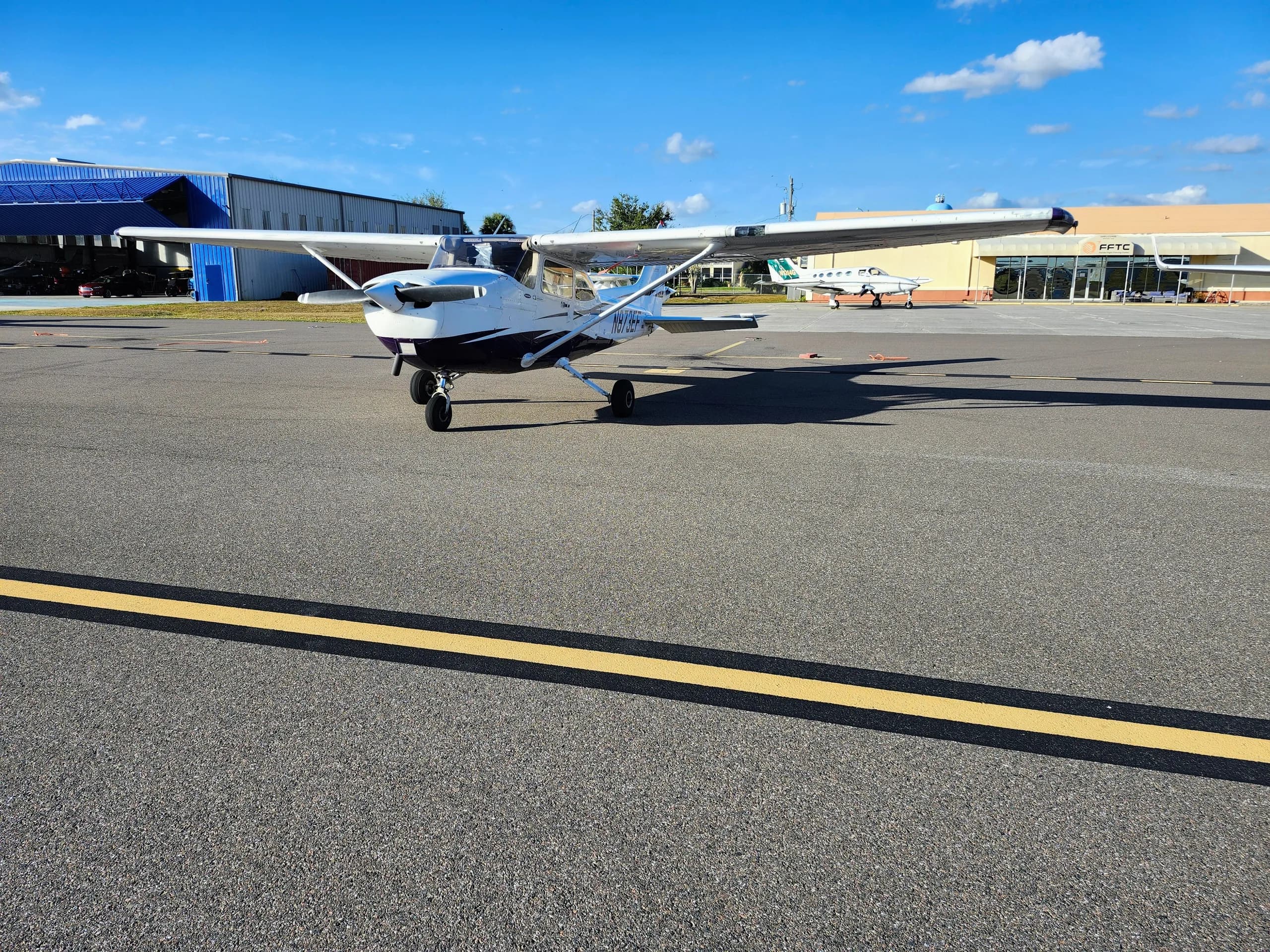 A white and blue Cessna 172, registration N873EF, parked on an airport tarmac with buildings in the background.